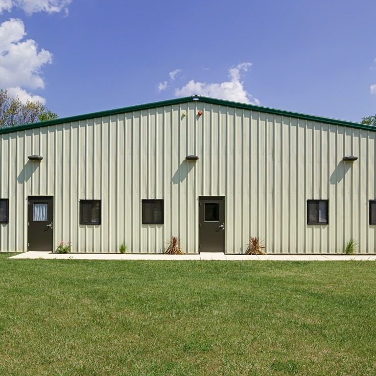 One-story metal building with green accents, featuring four doors and multiple windows, set in a grassy area under a blue sky.